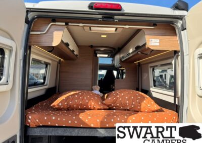 Rear interior of a camper van with a bed made up in orange polka-dot bedding and wood cabinetry above, daylight from windows on both sides, Swart Campers logo in corner.