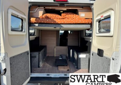 Open rear of camper van showing a raised bed with orange polka-dot bedding, wood storage cabinets, and grey upholstered storage bins.