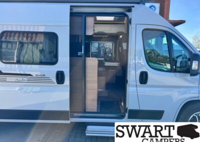 White camper van with slide-out door open, showing a compact interior with wooden cabinetry and a step leading inside, SWART CAMPERS logo overlay bottom right.