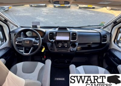 Interior view of a Fiat camper van cockpit: steering wheel, center touchscreen, gear selector, and dark dashboard with two front seats.