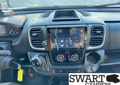 Center dashboard of a pickup truck showing a Pioneer touchscreen, round climate controls, gear shifter, cup holder, and keys with a yellow tag; Swart Campers logo bottom right.