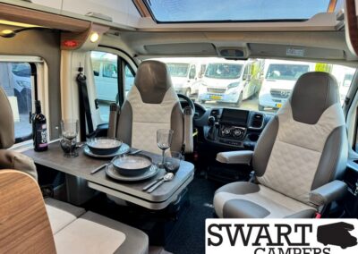 Interior of a camper van with a dining table set for two, plates, glasses, and a bottle of wine; front seats and dashboard visible in background.