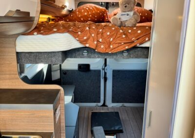 Cozy camper van bedroom with a raised bed, orange polka-dot bedding, and a teddy bear on the bed; kitchenette area to the left.