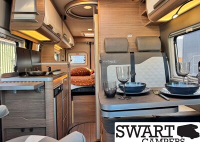 Modern camper van interior with kitchenette on the left, a dining table set for a meal, and a back bedroom with an orange blanket behind.