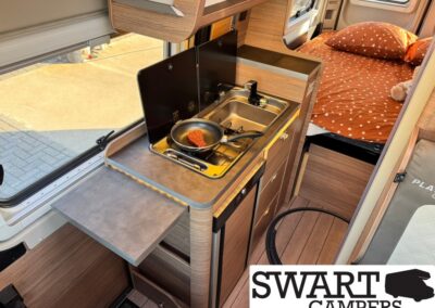 Compact camper van kitchen with a two-burner stove and sink, fold-out counter, and a bed with orange polka‑dot bedding in the background.