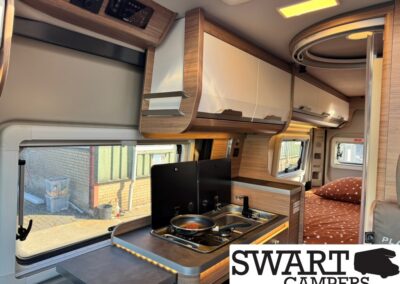 Interior of a modern camper van: compact kitchen with a two-burner stove and sink, wooden cabinets above, and a bed with orange dotted bedding visible in the back.