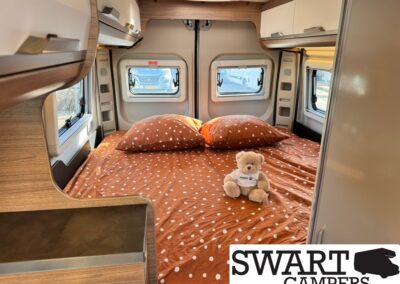 Camper van bedroom with brown polka-dot bedding, two pillows, and a teddy bear on the bed, surrounded by windows and wood cabinets.