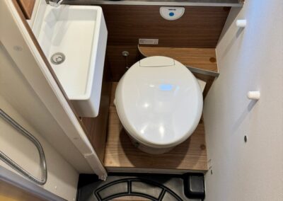 Compact camper bathroom with a white toilet on a wooden platform, a small sink to the left, and a toothbrush cup with brushes on the countertop