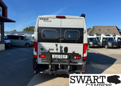 Rear view of a white KNAUS BoxLife camper with a black side stripe and two rear windows; bike rack attached below the bumper and a Dutch license plate KLK-05-P visible; SWART CAMPERS watermark in the corner.