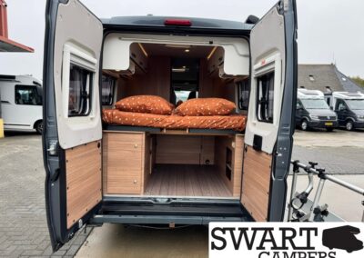 Rear view of a camper van with a raised bed and built-in wooden storage, orange polka-dot bedding, doors open.
