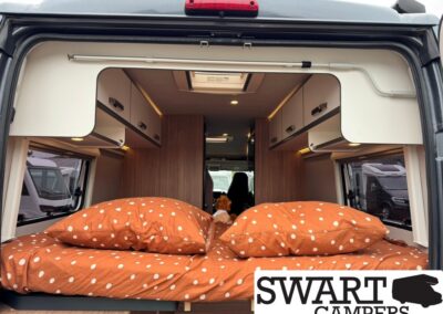 Rear view of a camper van interior with a bed made up in orange polka-dot bedding and overhead white cabinets above the sides.