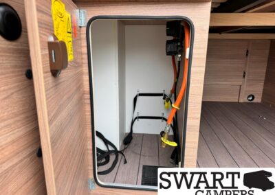 Open camper storage compartment with orange propane hose, gas regulator, and shutoff valve inside wood-paneled cabinet area
