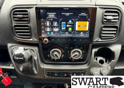 Center console of a vehicle showing a large touchscreen infotainment system (Android Auto) with climate control knobs below, gear shifter on the left, USB/12V outlets on the right, and a SWART CAMPERS logo in the bottom-right corner.