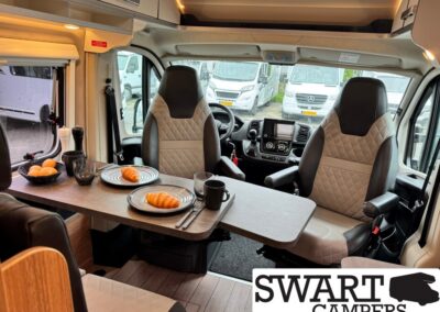 Camper van interior with a dining table set for two: croissants on plates, fruit bowl, mugs, and two captain chairs ready for breakfast.