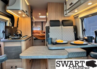 Interior of a camper van with a kitchenette, dining table set for two croissants, and a bed in the back with an orange blanket.