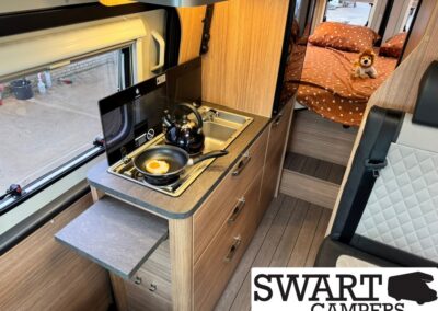 Camper van kitchen with a frying pan and egg on the stove, a kettle nearby, and wooden cabinets beside a bed with a stuffed lion toy in the background.