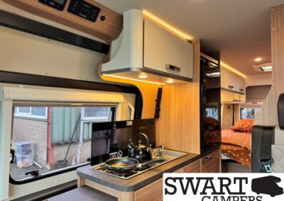 Interior of a modern camper van with a wood-toned kitchenette: two-burner stove, kettle, sink, and overhead/under-counter storage, with a bed in orange polka-dot bedding visible in the rear area.