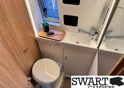 Compact camper bathroom with white toilet, sink, and mirror; toothbrush cup and brush on a wooden counter above storage cabinet.