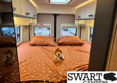 Camper van bedroom with orange polka-dot bedding, two pillows, and a lion plush on the bed under warm overhead lighting.