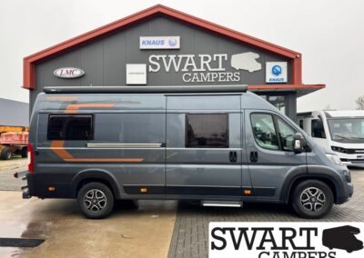 Gray camper van parked in front of a SWART CAMPERS storefront with a red roof and large logo sign above the entrance on a cloudy day, side window and roof rack visible.