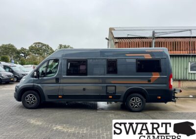 Side view of a dark gray camper van with orange accents and a solar panel on the roof, parked on a paved lot.